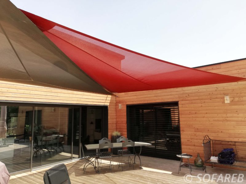Deux grandes voiles dombrage triangulaires marron et rouge accrochées au dessus de la terrasse dune maison en bois et fabriquée par Sofareb avec des tissus Serge Ferrari
