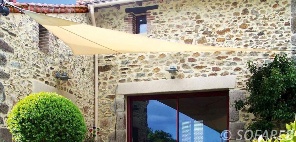 Voile ombrage sur-mesure beige adossée a la devanture d-une maison avec un revetement en pierre